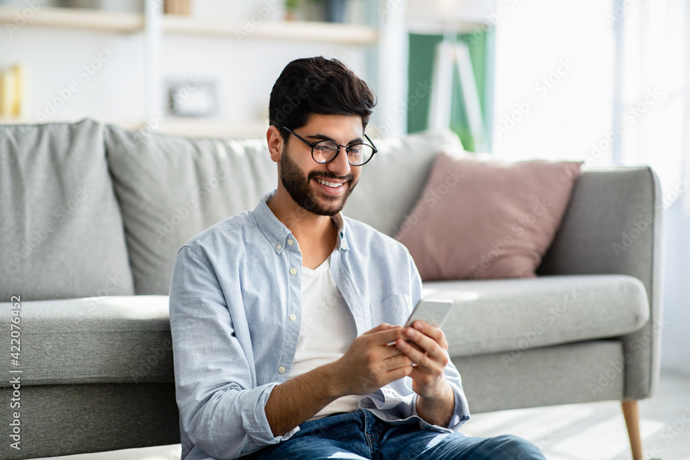 Arab guy texting and browsing internet on smartphone, resting at home, leaning on sofa Arab guy texting and browsing internet on smartphone, resting at home, leaning on sofa