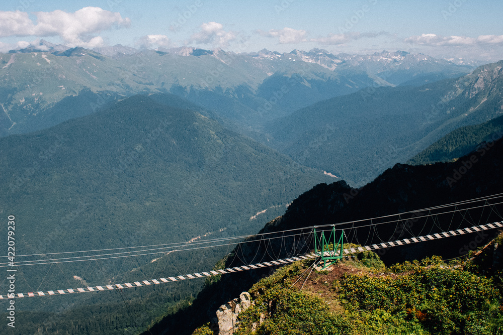 Rope bridge passing over an abyss in the mountains with visible clouds in the distance.