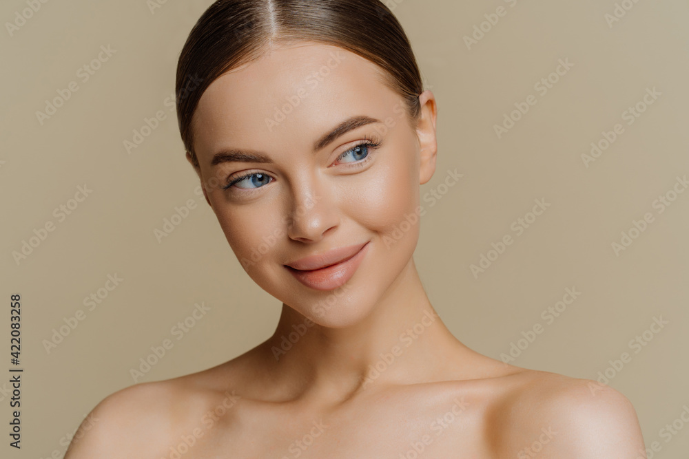 Portrait of thoughtful dreamy half naked woman looks aside with satisfied expression applies minimal makeup has fresh clean skin after facial beauty procedures isolated over beige background Portrait of thoughtful dreamy half naked woman looks aside with satisfied expression applies minimal makeup has fresh clean skin after facial beauty procedures isolated over beige background