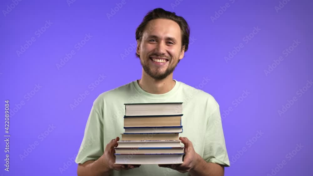 Male student holds stack of university books from library on violet background in studio. Guy smiles, he is happy to graduate. Male student holds stack of university books from library on violet background in studio. Guy smiles, he is happy to graduate.