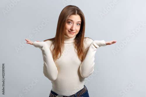 Portrait of puzzled cheerful girl showing don’t know gesture over isolated gray background. A young brown-haired woman in a white sweater spread her arms to the sides.