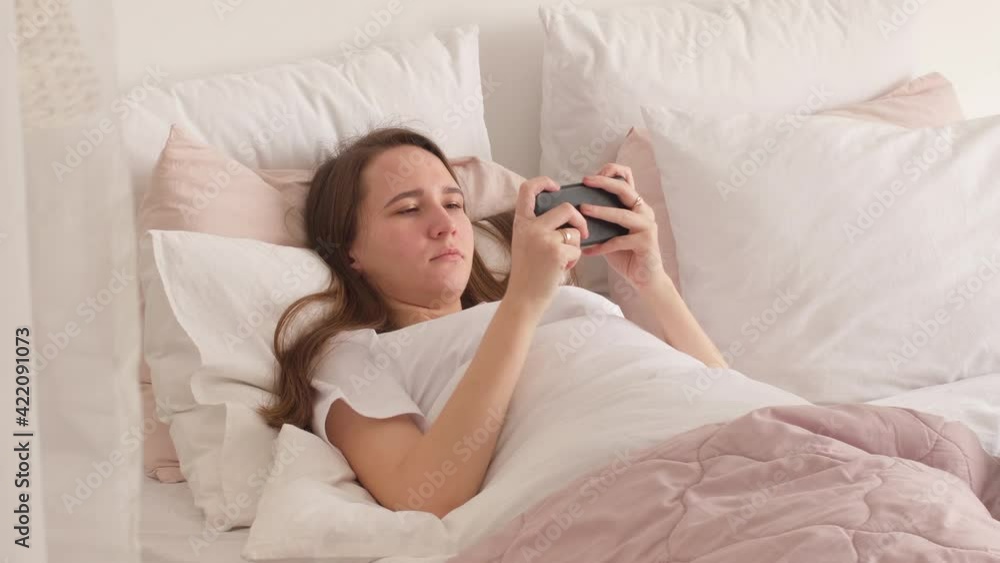 Beautiful girl lying on the bed in the room happily playing with her mobile phone. Young woman relax at home using smart phone Beautiful girl lying on the bed in the room happily playing with her mobile phone. Young woman relax at home using smart phone