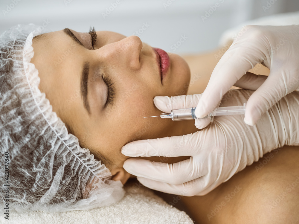 Naklejka premium Beautician doing beauty procedure with syringe to face of young brunette woman in sunny clinic. Cosmetic medicine and surgery, beauty injections