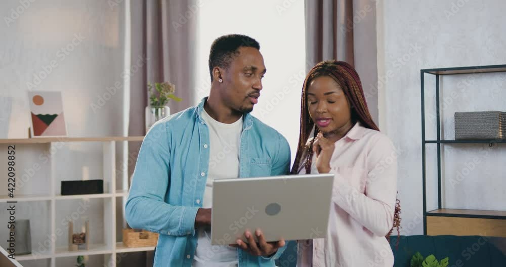Good-looking happy smiling young african american couple standing in front of camera and discussing creative ideas looking on laptop screen ,front view Good-looking happy smiling young african american couple standing in front of camera and discussing creative ideas looking on laptop screen ,front view