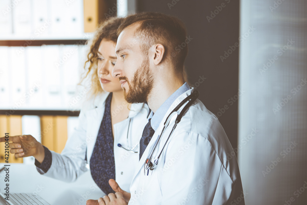 Red-bearded doctor with female colleague discussing current disease therapy while sitting at working place in sunny clinic. Team work in medicine Red-bearded doctor with female colleague discussing current disease therapy while sitting at working place in sunny clinic. Team work in medicine