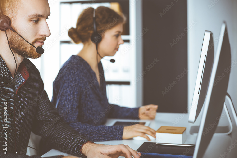 Red-bearded businessman talking by headset near his female colleague while sitting in sunny office. Diverse people group in call center Red-bearded businessman talking by headset near his female colleague while sitting in sunny office. Diverse people group in call center