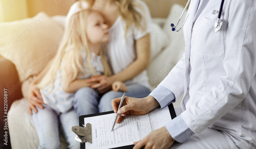 Fototapeta premium Doctor and patient. Pediatrician using clipboard while examining little girl with her mother at home. Sick and unhappy child at medical exam