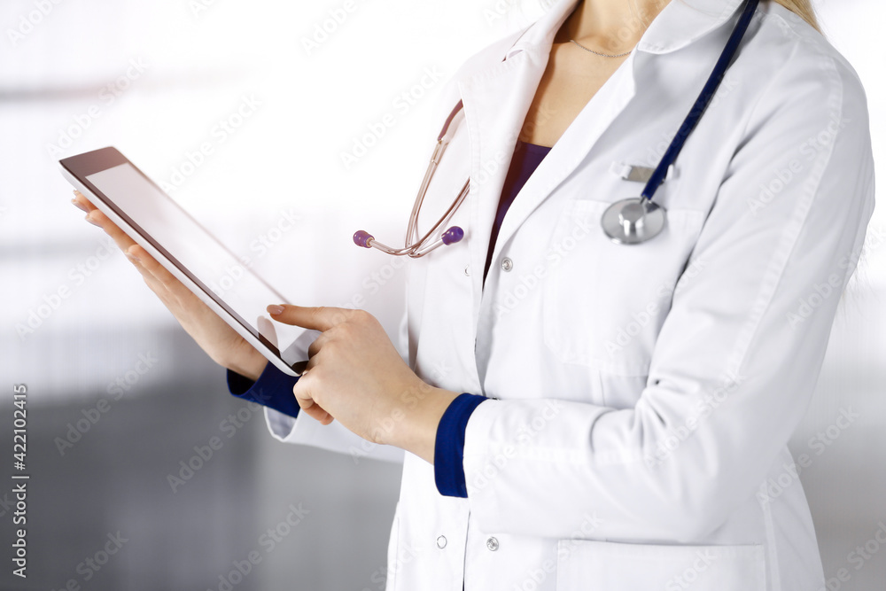 Unknown woman-doctor is holding a tablet computer in her hands, while standing in a clinic. Female physician with a stethoscope in her office, close-up. Perfect medical service in a hospital. Medicine