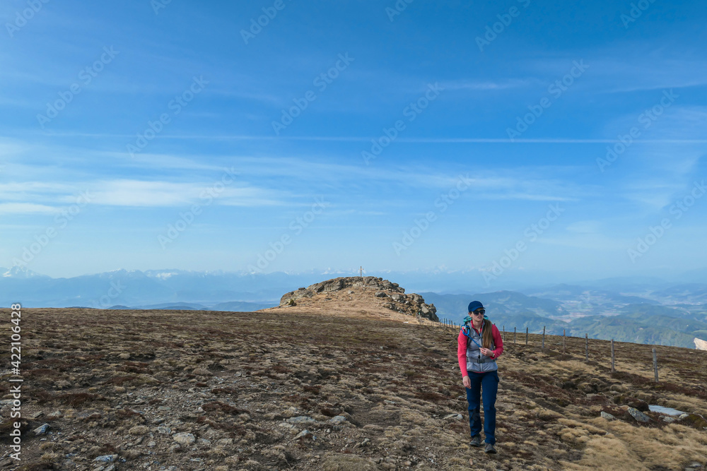 A woman with hiking backpack walking from the top of Sauofen in Austrian Alps. She is walking through a vast, golden pasture. Fall vibes. Mountain chains in the back. Serenity and achievement