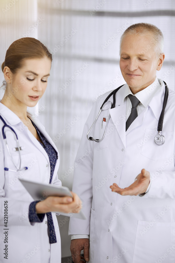 Elderly confident doctor and woman-physician discussing current disease therapy while standing in sunny clinic. Teamwork in medicine Elderly confident doctor and woman-physician discussing current disease therapy while standing in sunny clinic. Teamwork in medicine