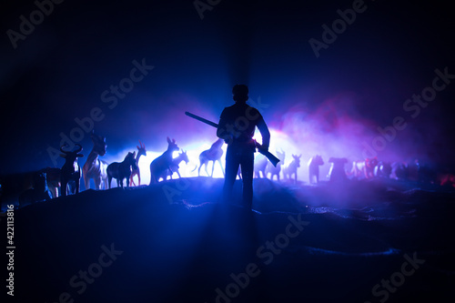 Silhouette of a man (hunter) with rifle standing against group of animals in colorful dark backlight. Decorated with miniatures. Selective focus