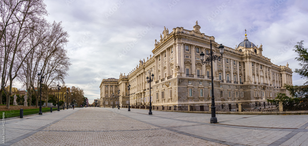 Side facade of the royal palace of Madrid, pedestrian street with lampposts, trees and sunny day with clouds.