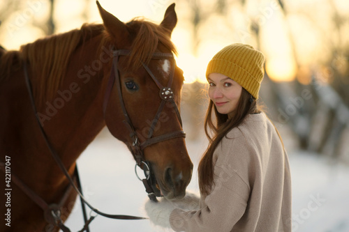 Side portrait of young woman and brown horse. Woman with long hear in yellow cap holding snaffle of horse