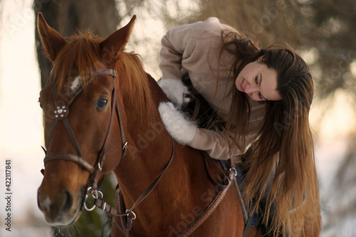 Close portrait of young woman with long hair in sweater sitting on brown horse at sunset. Background of winter trees