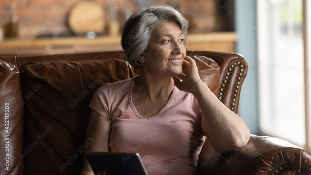 Nostalgia. Peaceful smiling elderly aged female grandma sit on comfy couch  hold tablet dream distracted from gadget screen. Pensive older hispanic  woman rest of pad computer lost in positive thoughts Stock Photo