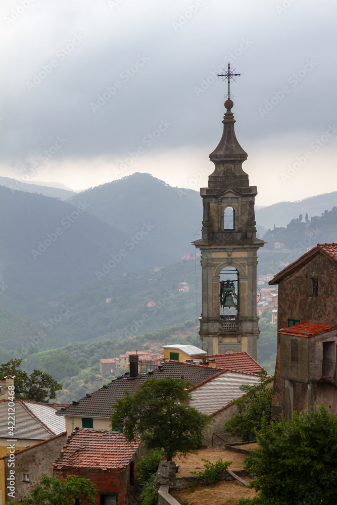 The bell tower of the temple against the background of mountains, surrounded by houses with tiled roofs. Low clouds after rain.