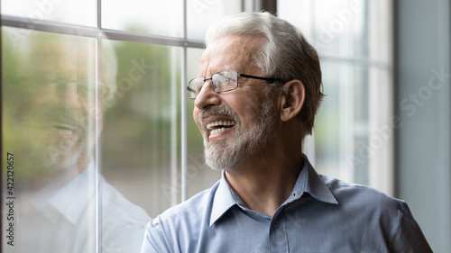 Optimistic dreamer. Energetic old man pensioner look at distance with healthy hollywood smile enjoy good day. Healthy aged male stand by window watch something in yard with candid laughter. Copy space