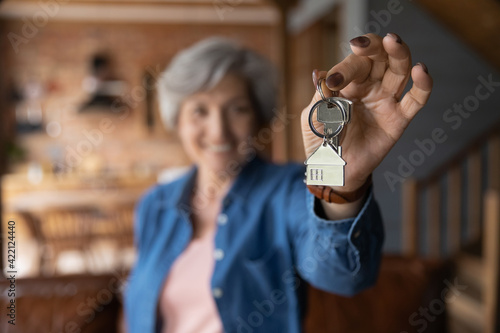 House of dream. Blurred portrait of happy elderly female of senior age becoming landlord homeowner pose in modern country cottage hold bunch of keys celebrate moving day. Focus on key in old lady hand
