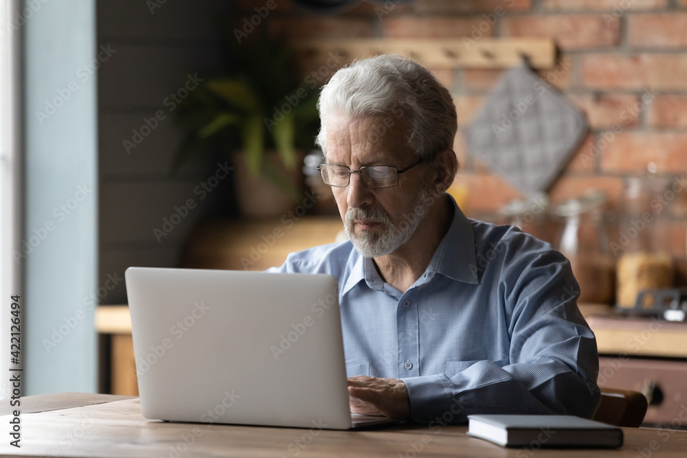 Aged remote worker. Concentrated senior male in glasses work on laptop from home office read email electronic document. Old age man employee freelancer sit at kitchen table by pc typing report online Aged remote worker. Concentrated senior male in glasses work on laptop from home office read email electronic document. Old age man employee freelancer sit at kitchen table by pc typing report online