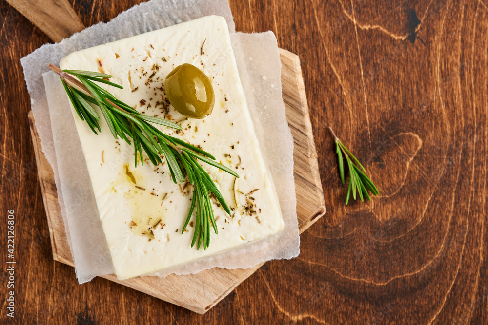 Cheese feta with rosemary, herbs, olives and olive oil on wooden cutting board on old wooden background. Traditional Greek homemade cheese. Selective focus. Cheese feta with rosemary, herbs, olives and olive oil on wooden cutting board on old wooden background. Traditional Greek homemade cheese. Selective focus.