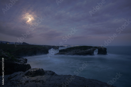 Full moon just before sunrise over the iconic arch of As Catedrais beach