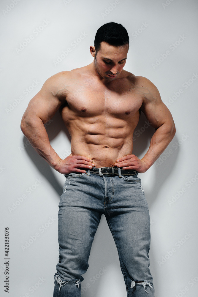 A young athlete bodybuilder poses in the studio topless in jeans near the wall. Sport. A young athlete bodybuilder poses in the studio topless in jeans near the wall. Sport.