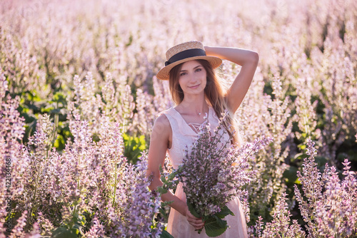 Young blonde woman in straw hat on the background of blooming field of pink sage. Close-up portrait beautiful girl holding bouquet flowers in hands. Weekend walk outside the city. Agricultural texture