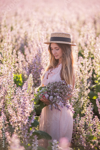 Young blonde woman in straw hat looks into the distance against the background of blooming field pink sage. Close-up portrait of beautiful girl holding bouquet flowers in hands. Agricultural texture