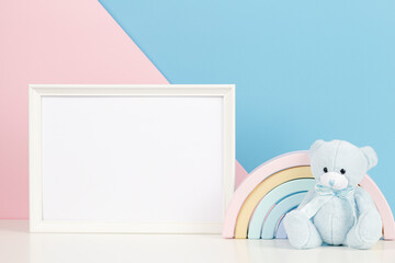White blank frame with teddy bear and pastel toy rainbow on white desk. Baby room art frame mock up with baby kid toys over pink and blue background
