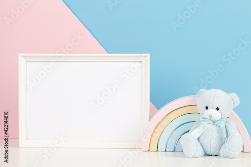 White blank frame with teddy bear and pastel toy rainbow on white desk. Baby room art frame mock up with baby kid toys over pink and blue background © vejaa