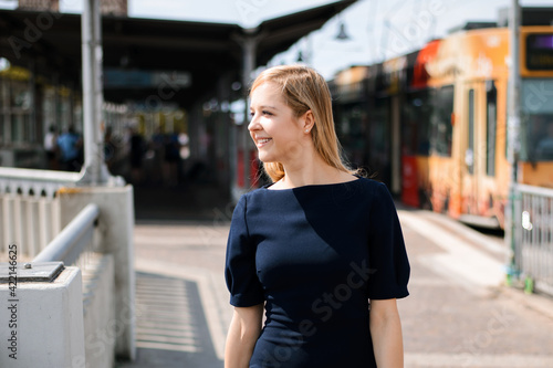 Portrait of young blond woman with toothy smile standing at train station and looking to the side. Cheerful girl in dark blue dress in the vacation.