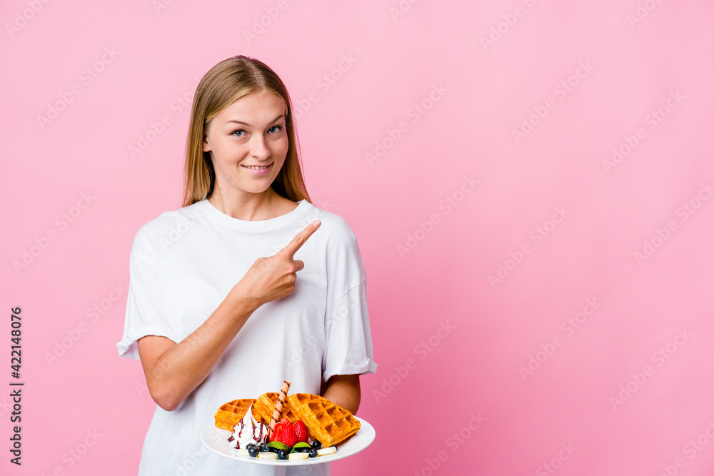 Young russian woman eating a waffle isolated smiling and pointing aside, showing something at blank space. Young russian woman eating a waffle isolated smiling and pointing aside, showing something at blank space.