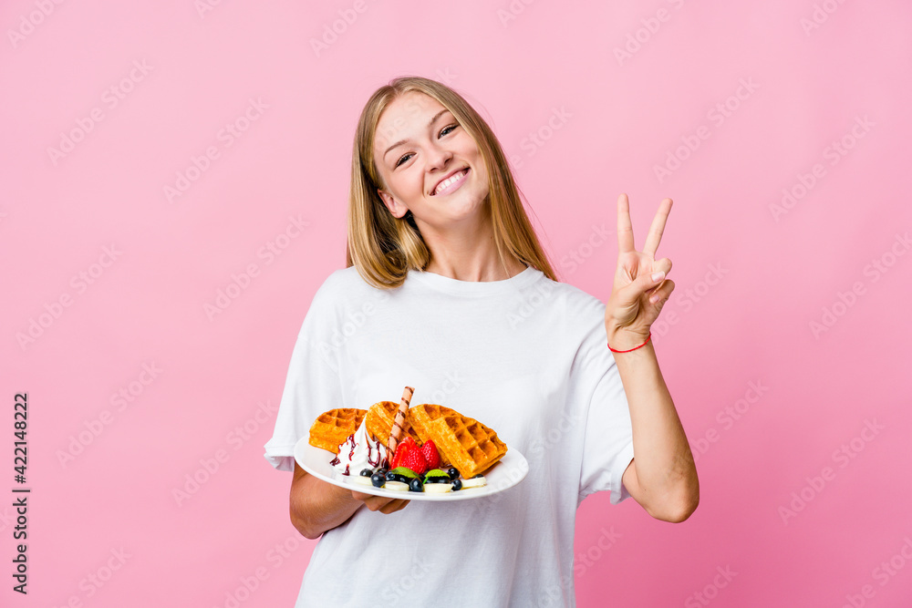 Young russian woman eating a waffle isolated joyful and carefree showing a peace symbol with fingers. Young russian woman eating a waffle isolated joyful and carefree showing a peace symbol with fingers.