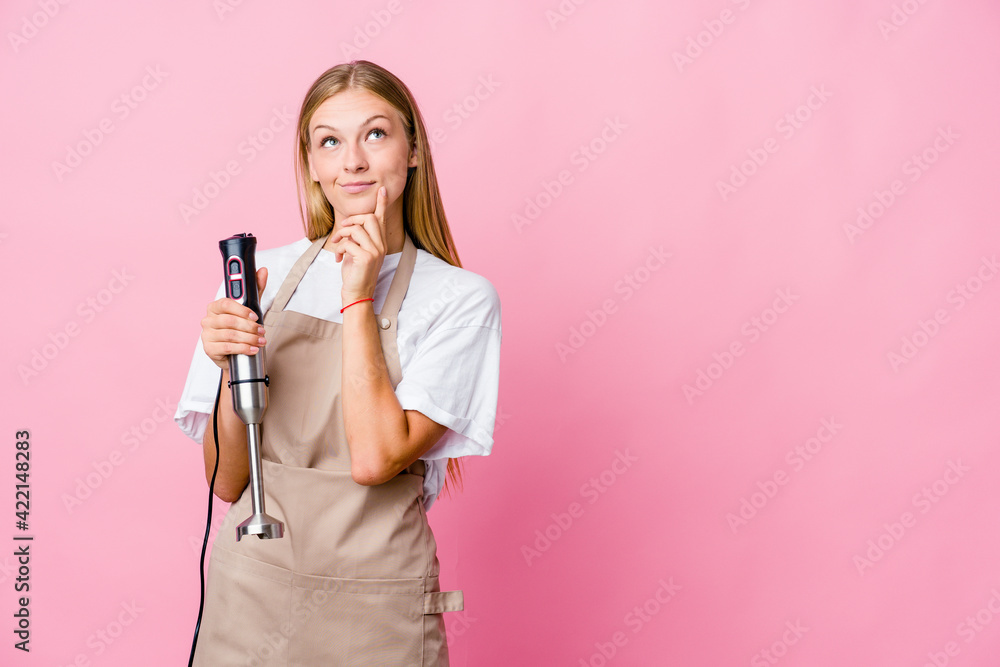 Young russian cook woman holding an electric mixer isolated looking sideways with doubtful and skeptical expression. Young russian cook woman holding an electric mixer isolated looking sideways with doubtful and skeptical expression.