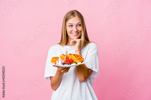 Young russian woman eating a waffle isolated keeps hands under chin, is looking happily aside.