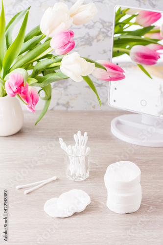 Personal hygiene, cleanliness and skin care. Cotton pads and swabs in a glass on a table in front of a mirror and a bouquet of tulips in a vase. Vertical view