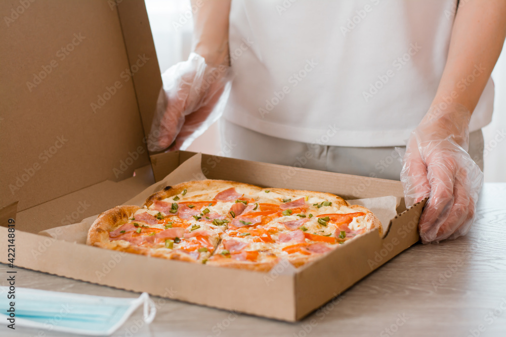 Takeaway food. A woman in disposable gloves holds a cardboard box of pizza and a protective mask on the table in the kitchen