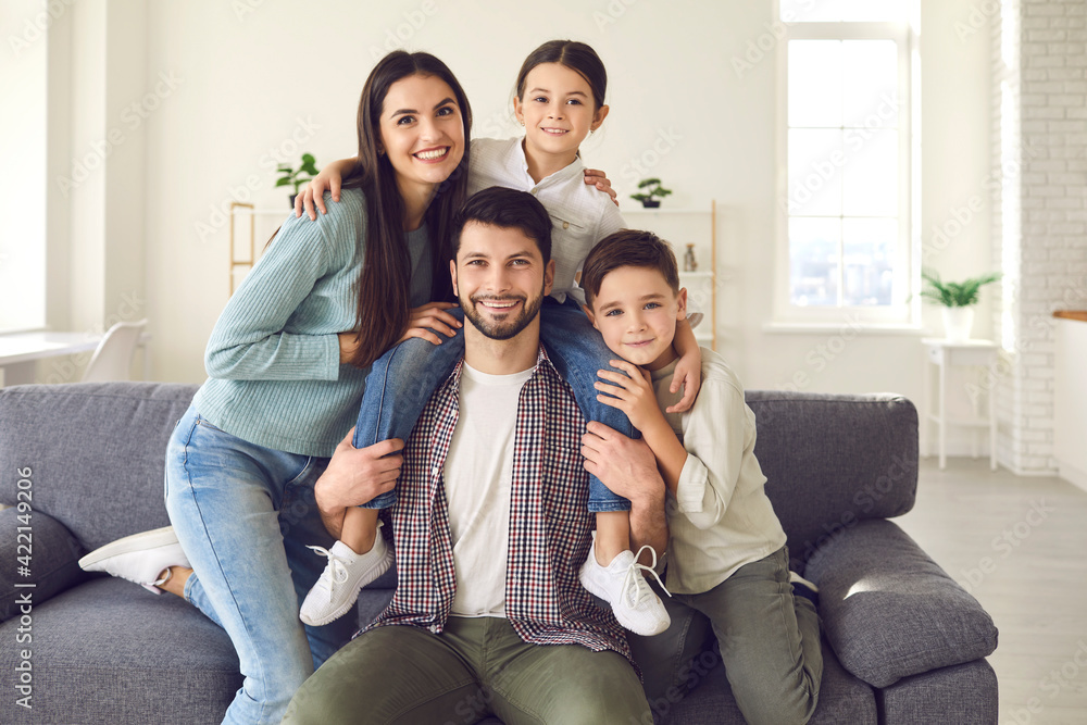 Portrait of smiling mother, father and children at home. Happy young couple with kids sitting on sofa in the living-room of their new house or apartment and looking at camera during family video call