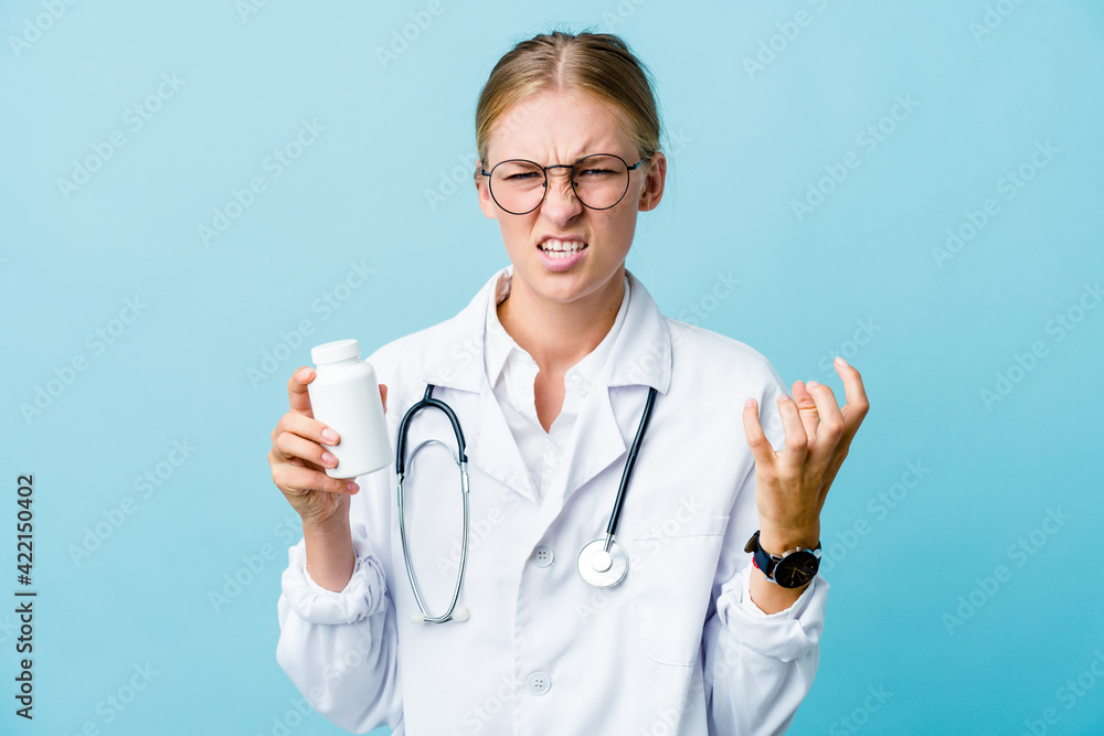 Young russian doctor woman holding pills bottle on blue upset screaming with tense hands. Young russian doctor woman holding pills bottle on blue upset screaming with tense hands.