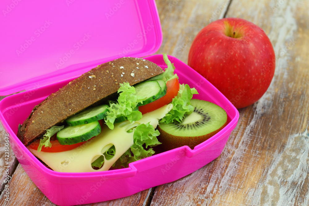 Whole grain roll with cheese, lettuce, tomato and cucumber, kiwi fruit and red apple in pink lunch box on rustic surface, closeup