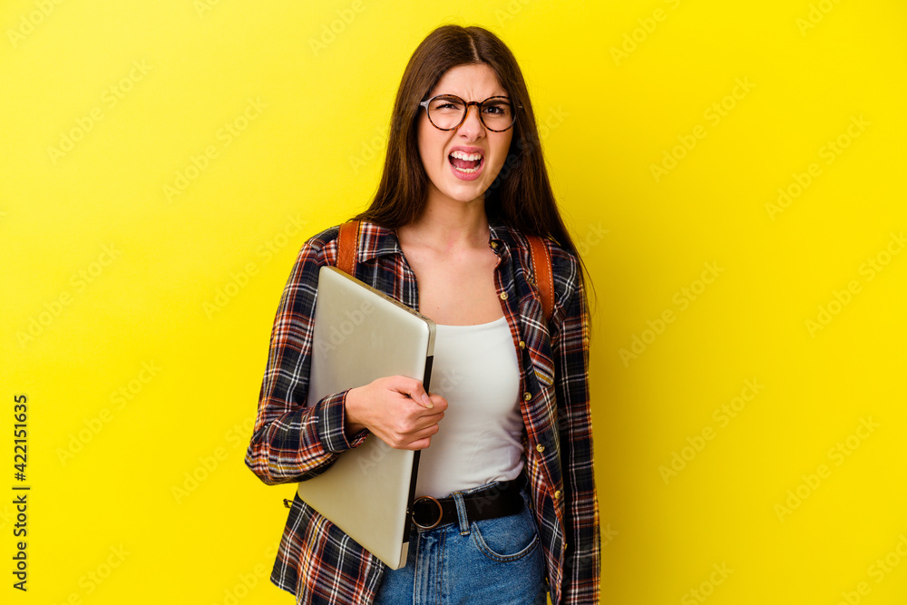 Young caucasian student woman holding a laptop isolated on pink background screaming very angry and aggressive. Young caucasian student woman holding a laptop isolated on pink background screaming very angry and aggressive.