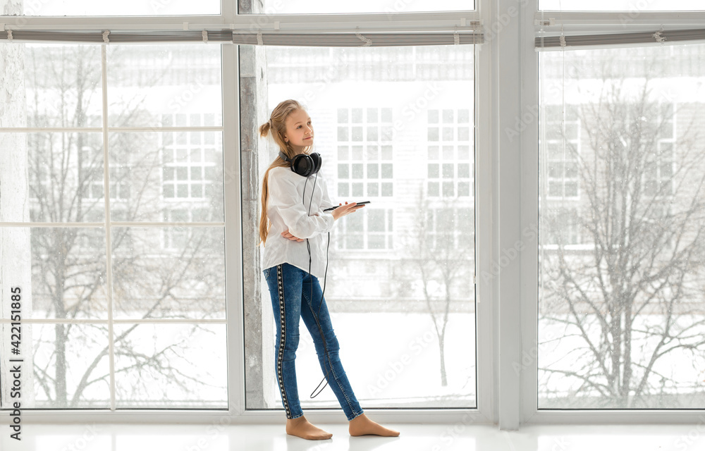 A girl on the windowsill by a huge window turns on a playlist on her smartphone. The child enjoys music in large headphones. A girl on the windowsill by a huge window turns on a playlist on her smartphone. The child enjoys music in large headphones.