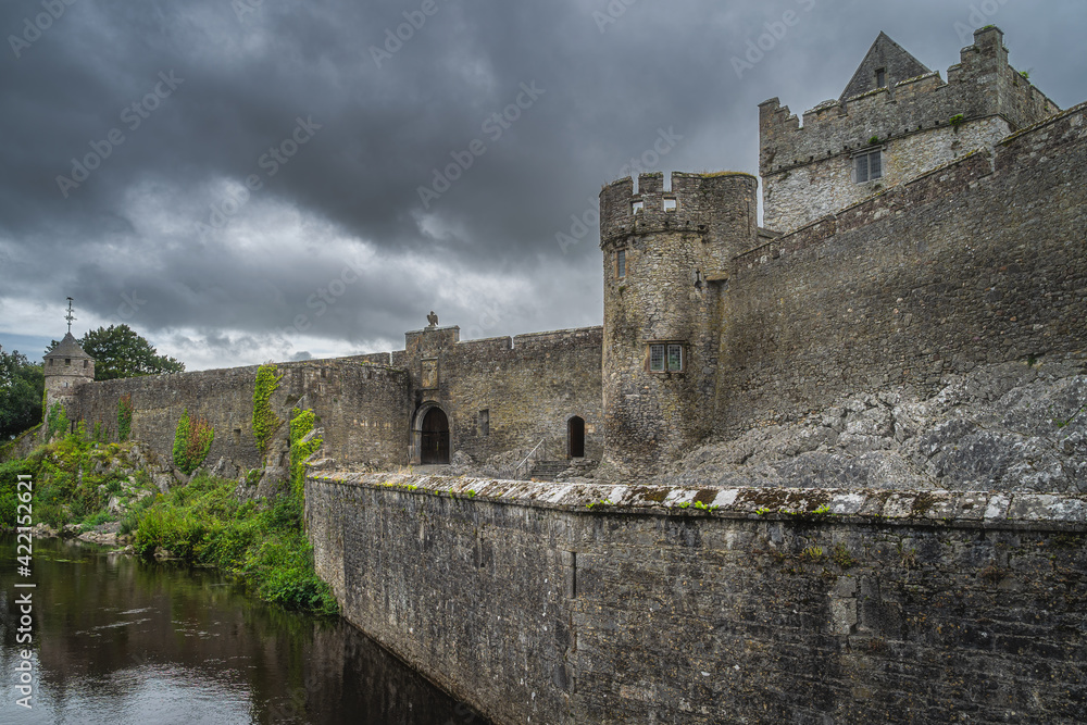 Tall stone walls and moat of 11th century Cahir castle in Cahir town with dramatic, storm sky in background, County Tipperary, Ireland