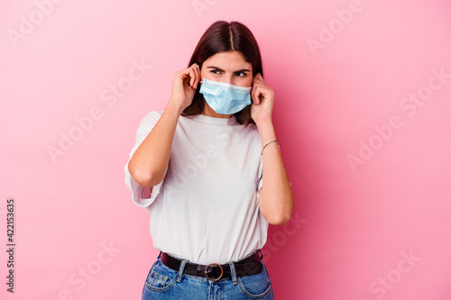 Young caucasian woman wearing a mask for virus isolated on pink background covering ears with hands.