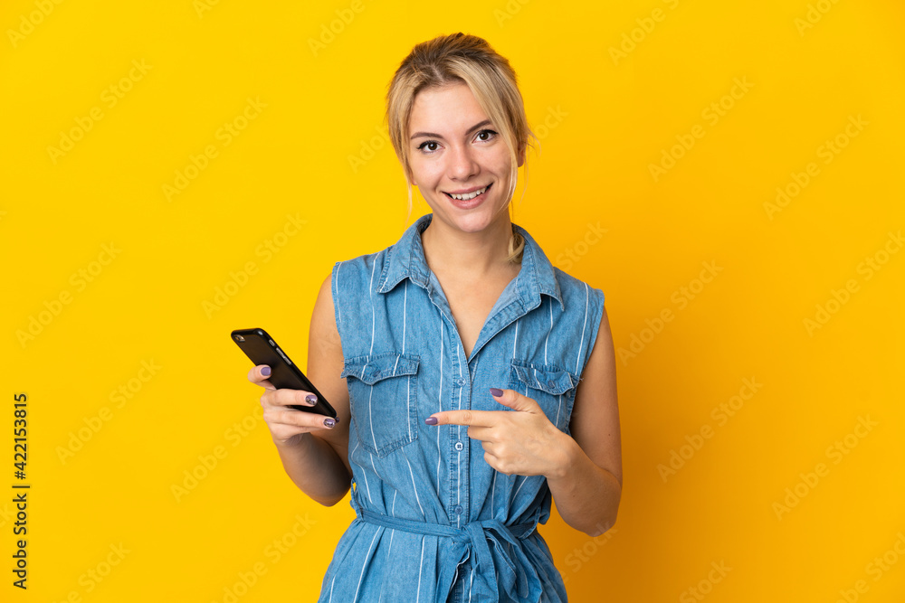 Young Russian woman isolated on yellow background using mobile phone and pointing it Young Russian woman isolated on yellow background using mobile phone and pointing it