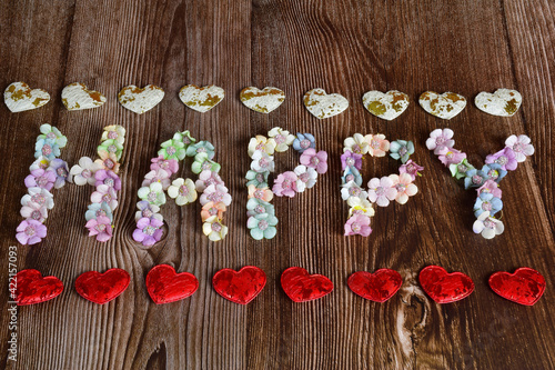 On a wooden background  the word HAPPY is spelled out in large letters of multicolored flowers. Decorative hearts are lined top and bottom in a line