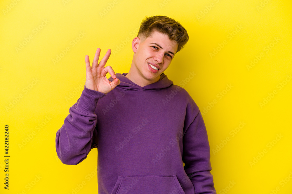 Young caucasian man isolated on yellow background winks an eye and holds an okay gesture with hand. Young caucasian man isolated on yellow background winks an eye and holds an okay gesture with hand.