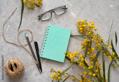 Elegant sunny composition with sprigs of mimosa, notebook, pen, pencil, glasses and skein of thread. Spring, joyful and happy mood. Spring renewal in nature. Still life with flowers. Grey background.