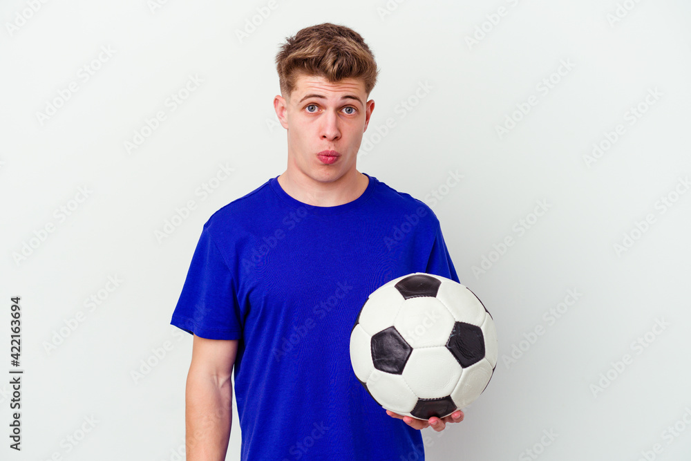 Young caucasian man playing soccer isolated on background shrugs shoulders and open eyes confused.