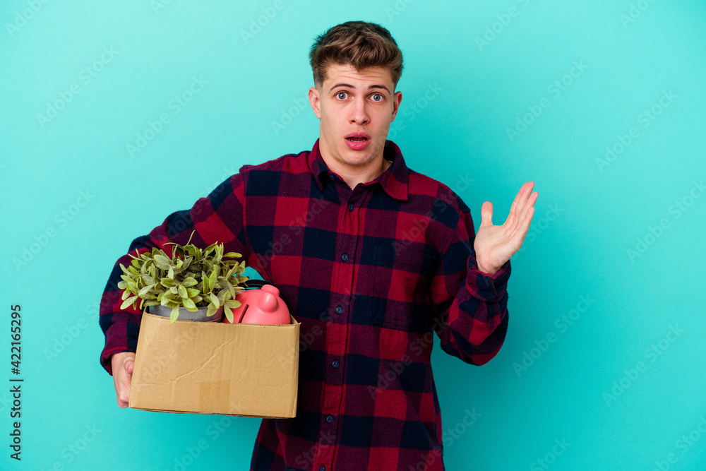 Young caucasian man moving holding a box isolated on blue background surprised and shocked.